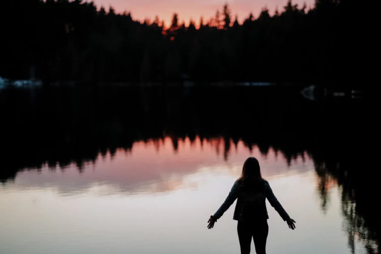 a woman standing by a lake with arms open looking at the sun setting, signifying the question how to let go and surrender