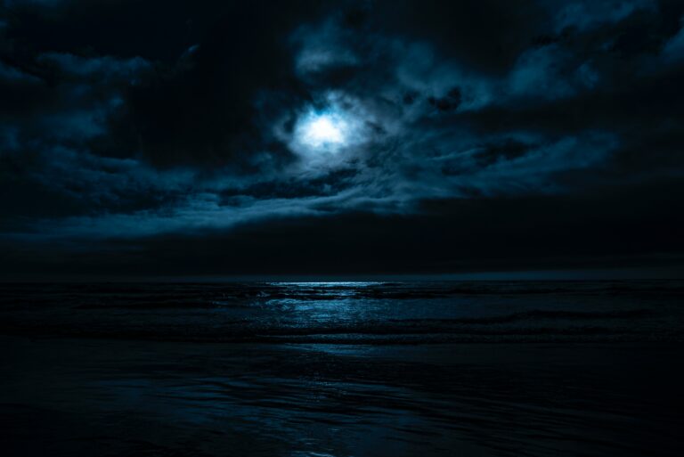 a dark night with a bright full moon behind clouds and an ocean visible representing dark night of the soul